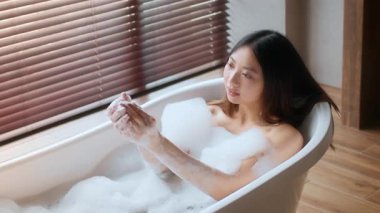 Şık bir banyoda, genç bir kadın kabarcıklarla dolu bir küvette rahatlar, kendi kendine bakım ritüeli yapar. Yumuşak ışık, ahşap panjurların arasından süzülür ve sakin atmosferi güçlendirir..