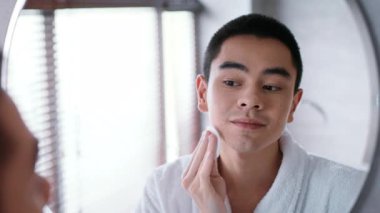Çağdaş bir banyoda, genç bir Asyalı aynaya bakarken cilt bakım ürünü uyguluyor. Rahat bir durumda, günlük şımartma rutini sırasında bornoz giyiyor..