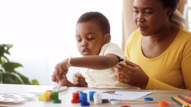 Genç bir Afrikalı Amerikalı çocuk masada otururken canlı renklerle resim yapıyor. Sevgi dolu bir yetişkin ona arkadan destek olur, yaratıcılığı teşvik eder ve ev ortamında bağ kurar..