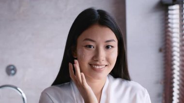 In a modern bathroom, a young Asian woman applies skincare on her face while smiling radiantly. Her routine reflects a commitment to self-care and beauty in her daily life.