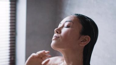 Sakin bir banyoda, genç bir Asyalı kadın buharın rahatlatıcı sıcaklığının tadını çıkarırken günlük cilt bakımını yapıyor. Şımartma ayinlerinin ortasında rahatlamış ve tazelenmiş görünüyor..