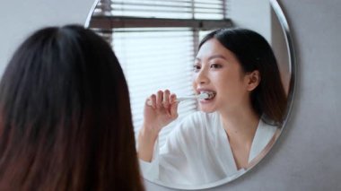 Genç bir kadın günlük kişisel bakım rutiniyle meşgul, aynaya bakarken dişlerini fırçalıyor. Sıcak banyo atmosferi gevşemeyi ve sağlığı teşvik ediyor..