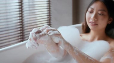 Sakinleştirici bir banyoda, genç bir Asyalı kadın şımartıcı bir köpük banyosunun keyfini çıkarıyor. Ellerine sabunla nazikçe masaj yapar, özgüven rutinini ve rahatlığını benimser..