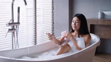 Şık bir banyoda, genç bir Asyalı kadın köpük banyosu yaparken şımartıcı bir cilt bakımına dalar. Yumuşak ışık, kişisel bakım için sakin bir atmosfer yaratır..