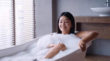 Genç Asyalı bir kadın banyo köpükleriyle dolu bir küvette rahatlıyor ve modern bir banyoda vücut bakımını sergiliyor. Cildini şımartırken ve huzurun tadını çıkarırken mutlu görünüyor..