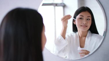 Sıcak bir banyo ortamında, genç bir Asyalı kadın yansımasına gülümserken yüzüne serum uyguluyor. Rahatlamak ve şımartmak için günlük cilt bakımı ritüelini benimsiyor..
