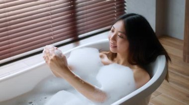 Genç bir Asyalı kadın huzurlu bir banyoda kabarcıklarla dolu bir küvette rahatça oturuyor. Güneş ışığı, ahşap panjurların arasından süzülüp şımartıcı rutinini arttırıyor..