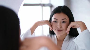 İyi aydınlatılmış bir banyoda, genç bir Asyalı kadın aynaya bakarken cilt bakımını yapar. Yüzüne nazikçe masaj yapar, rahatlama ve kendine bakmanın önemini vurgular..