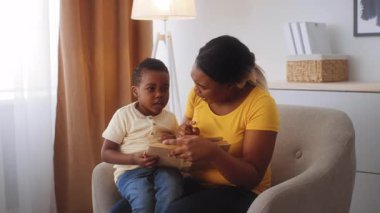 Bir anne ve küçük oğlu evde hassas bir anı paylaşıyorlar. Çocuk, sevgi dolu bir şekilde kucağına oturur ve onunla sıcak, rahat bir yaşam alanında iletişim kurar..