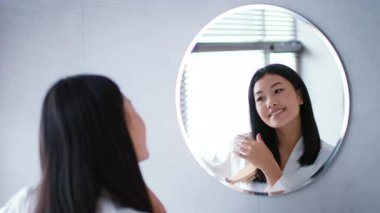 Parlak bir banyoda, genç bir Asyalı kadın saçlarını tararken, saçlarına bakım ürünleri sürerken gülümsüyor ve saçlarını tarıyor.