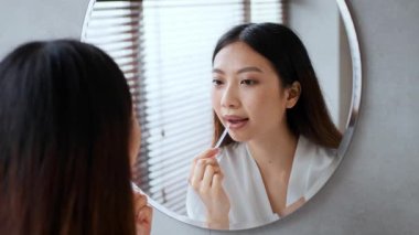 Genç bir kadın şık bir banyoda deri diyetine odaklanır. Etrafı rahatlama ve şımartma atmosferiyle çevrili yuvarlak bir aynaya bakarken makyaj yapar..