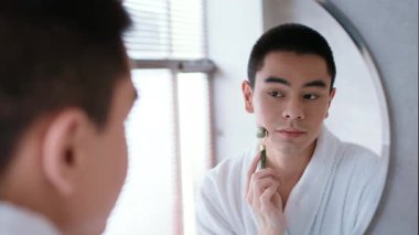 Beyaz bornozlu genç bir Asyalı, parlak, modern bir banyoda aynadaki yansımasına bakarken yüzünde bir silindir ile cilt bakımı rutinini yapar..