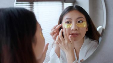 Sakin bir banyoda, genç bir Asyalı kadın aynaya bakarken göz altı bantları uyguluyor. Bu an, kendini özgüvene ve evde şımartmaya olan bağlılığını yansıtıyor. Rahatlatıcı bir rutini kucaklıyor..