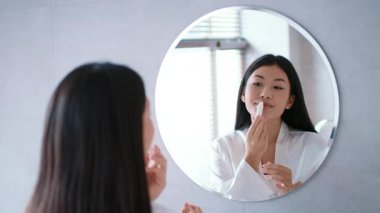 Genç bir Asyalı kadın kendini şımartırken dudak bakım ürünlerini banyoda uyguluyor. Temiz, modern alan kendine bakım ritüelini geliştiriyor. Doğal ışık alanı dolduruyor..