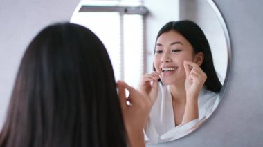 Çağdaş bir banyoda, genç bir Asyalı kadın şımartıcı rutinini uyguluyor, evde dişlerini temizlerken gülümsüyor, rahatlatıcı bir atmosfer yaratıyor..