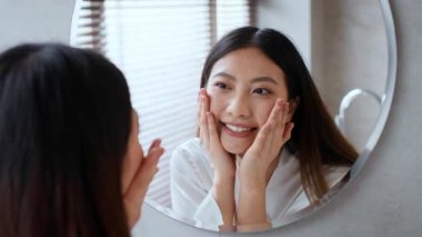 Genç bir Asyalı kadın, aynadaki yansımasına gülümserken cilt ürünlerini uygulayarak banyoda kendine bakıcılık yapıyor. Bu an, rahatlamanın ve şımartmanın özünü yakalar..