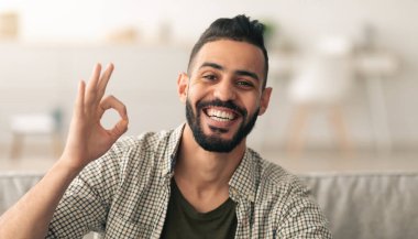 Sakallı bir adam el hareketi yaparken geniş bir gülümseme sergiliyor. İçerde rahat bir konumda oturuyor. Etrafı yumuşak ışıkla dolu rahat bir oturma odası atmosferiyle çevrili..