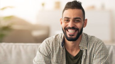 Sakallı bir adam kanepede oturur, sıcak bir şekilde gülümser. Oturma odasında yumuşak bir ışık ve rahat bir atmosfer var. Çevresinde neşeli ve rahat görünüyor..