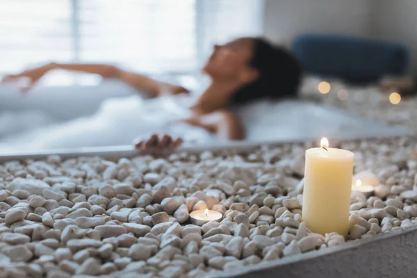 Sakin bir spa deneyimi, pürüzsüz taşlarla çevrili bir banyoda dinlenen bir kadın içerir. Yumuşak mum ışığı, sakin atmosferi güçlendirir, barışı ve rahatlığı teşvik eder..