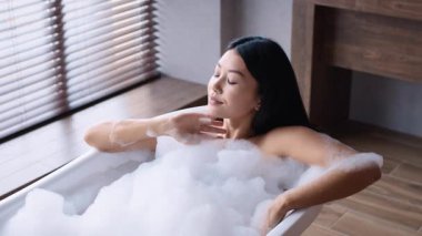 Genç Asyalı bir kadın evinde rahatlatıcı bir köpük banyosunun keyfini çıkarıyor, doğal ışıkla dolu şık bir banyoda günlük cilt bakımını yapıyor, rahatlatıcı bir ortam yaratıyor..