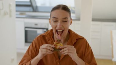 Bir kadın elinde bir dilim pizza tutarak evinde mutlu bir şekilde oturuyor. Sıcak ortamında büyük bir ısırık alır, rahatlığı ve keyfi yansıtır..