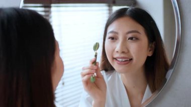 Genç bir Asyalı kadın banyoda güne hazırlanıyor. Yüzüne yeşim taşı takarken gülümsüyor. Yumuşak ışık pencereden süzülüyor, sakin özgüven anlarını geliştiriyor..