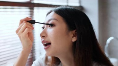 Genç bir Asyalı kadın iyi aydınlatılmış bir banyoya rimel sürüyor. Sakinleştirici bir atmosferle çevrili kişisel bir kişisel bakım rutinine daldı. Yumuşak ışık onun odaklanmış ifadesini arttırır..