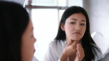 A young woman examines her skin closely in a bathroom mirror, engaging in a personal care routine. She appears attentive as she checks for blemishes and applies skin care treatments.