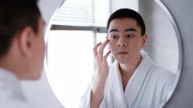 Çağdaş bir banyoda, genç bir Asyalı aynaya bakarken cilt ürünleri uyguluyor. Bu günlük kişisel bakım ritüeli yeni bir ışıltı arayan yeni nesil için vücut bakımını vurguluyor..