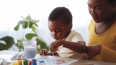 Doğal ışıkla dolu parlak bir odada, genç bir Afrikalı Amerikalı çocuk annesiyle birlikte resim yapıyor, yaratıcılık yoluyla bağlarını sergiliyor ve sanatsal ifadedeki mutluluğu paylaşıyor..