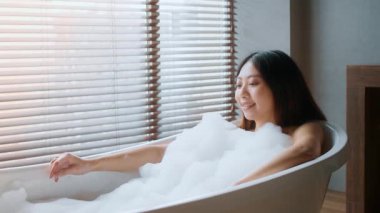 Genç bir Asyalı kadın banyoda kabarcıklarla dolu şımartıcı bir küvetin keyfini çıkarıyor. Güneş ışığı pencereden süzülerek rahatlatıcı bir atmosfer yaratıyor..