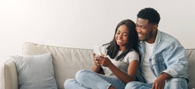 Birlikte vakit geçirmek. Akıllı telefonuyla sosyal medyayı paylaşan mutlu zenci kadın portresi gülümseyen erkek arkadaşıyla koltukta oturan çift, evdeki oturma odasında dinlenen iki arkadaş.