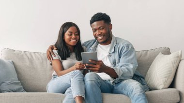 Hafta sonu eğlencesi. Evlerinde tablet kullanan, internete giren veya ne alacaklarını seçen, oturma odasındaki rahat koltukta oturan mutlu ve sevecen genç siyah çift. Kadını kucaklayan adam, bedava kopya alanı.