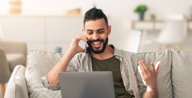 Bir adam koltukta otururken telefonla konuşmaktan mutlu olur. Dizüstü bilgisayarını kullanıyor. Etrafı parlak güneş ışığıyla çevrili oturma odasıyla çevrili..