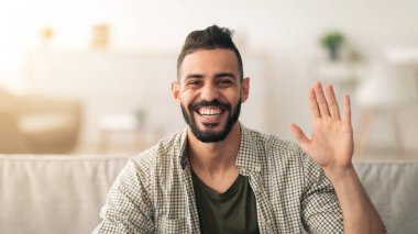 Sakallı ve şık saçlı bir adam kanepeye oturup elini sallarken gülümsüyor. Işıl ışıl, rahat oturma odasında yumuşak mobilyalar ve sıcak ışıklandırma var..