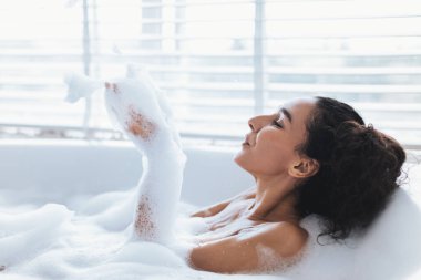 Köpük dolu bir küvette oturan bir kadın köpükle oynarken gülümsüyor. Doğal ışık, pencere perdelerinden süzülerek rahatlatıcı ve sakin bir atmosfer yaratır..