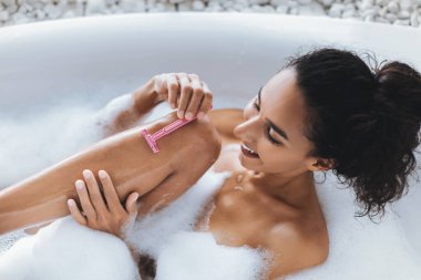 Sakin bir banyo ortamında kıvırcık saçlı bir kadın, bacağını nazikçe tıraş ederken köpük banyosundan zevk alır. Sıcak su ve sabun köpükleri rahatlatıcı bir atmosfer yaratır..