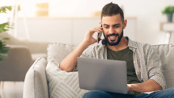 Bir adam kanepeye rahatça oturur, dizüstü bilgisayarında yazarken telefonla konuşur. Parlak, havadar oturma odası bitkiler ve yumuşak mobilyalarla dekore edilmiştir..