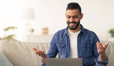 A man with a beard smiles and gestures while participating in a virtual meeting. He sits on a sofa with a laptop, enjoying a relaxed atmosphere at home during the day.