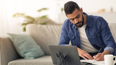 Genç bir adam evdeki işine odaklanıyor, not defterine notlar yazıyor. Önünde bir dizüstü bilgisayarla koltukta rahatça oturuyor. Oda aydınlık ve davetkar..