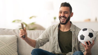 Neşeli bir adam bir elinde futbol topu diğer elinde uzaktan kumandayla evdeki bir kanepede oturur. Heyecanlı, ekrandaki bir futbol maçından zevk alıyor..