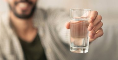 Bir adam su dolu şeffaf bir bardak tutar, birine sunarken sıcak bir gülümsemeyle. Arka plan hafifçe bulanık, rahatlatıcı bir atmosfer yaratıyor..