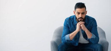 Gri bir sandalyede oturan sakallı bir adam dalgın görünüyor. Ellerini birbirine kenetledi ve düşüncelerde kaybolmuş gibi görünüyor. Çevresi asgari ve hafif aydınlık..