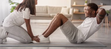 Ev antrenmanı. Genç Afrikalı Amerikalı çift oturma odasında birlikte egzersiz yapıyor, siyah kız arkadaşına gülümseyen erkek arkadaşına karın kaslarını çalıştırması için yardım ediyor, bacakları ve ayakları tutuyor, profil manzarası...