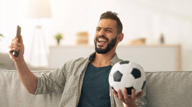 Bir adam televizyonda futbol maçı izlerken sevinçle kutlama yapar. Bir elinde futbol topu, diğer elinde uzaktan kumandayla oturma odasının rahatlığından heyecanını gösteriyor..