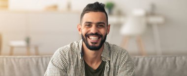 Neşeli bir adam iyi aydınlatılmış bir oturma odasındaki bir kanepede rahatça oturur. Onun uzun bir sakalı var ve günlük bir gömlek giyiyor. Ortam rahat, yumuşak ışık ve modern dekorla..