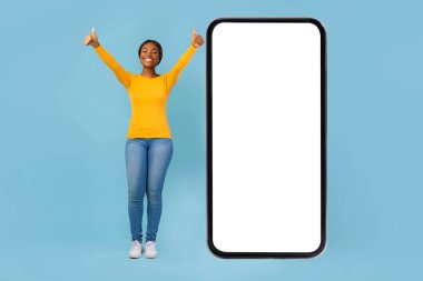 Genç bir kadın büyük bir gülümseme ile kendine güvenerek ayakta duruyor ve parmakları kocaman boş bir akıllı telefonun yanında. Arka plan parlak mavi, neşeli bir atmosfer yaratıyor..