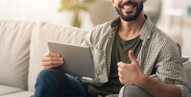 Sakallı bir adam elinde tablet tutarken gülümsüyor ve başparmağını kaldırıyor. Parlak ve rahat bir oturma odasında, bitkilerle ve doğal ışıkla bir kanepede oturuyor..