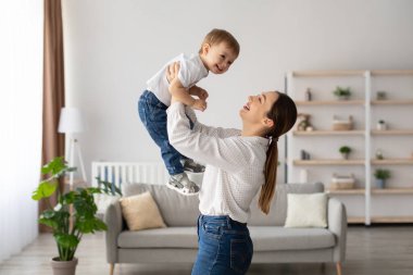 Neşeli bir anne gülümseyen bebeğini güneşli bir oturma odasında havada tutuyor. Mekan yumuşak renkler ve bitkilerle aydınlık, sıcak bir aile ortamı yaratıyor..