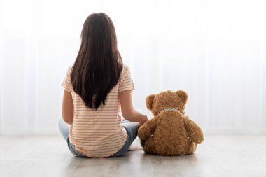 Bir kız yerde oturuyor, sırtı seyirciye dönük, oyuncak ayının yanında. Parlak bir pencereden dışarı bakıyorlar, rahat bir yerde huzurlu bir anın tadını çıkarıyorlar..